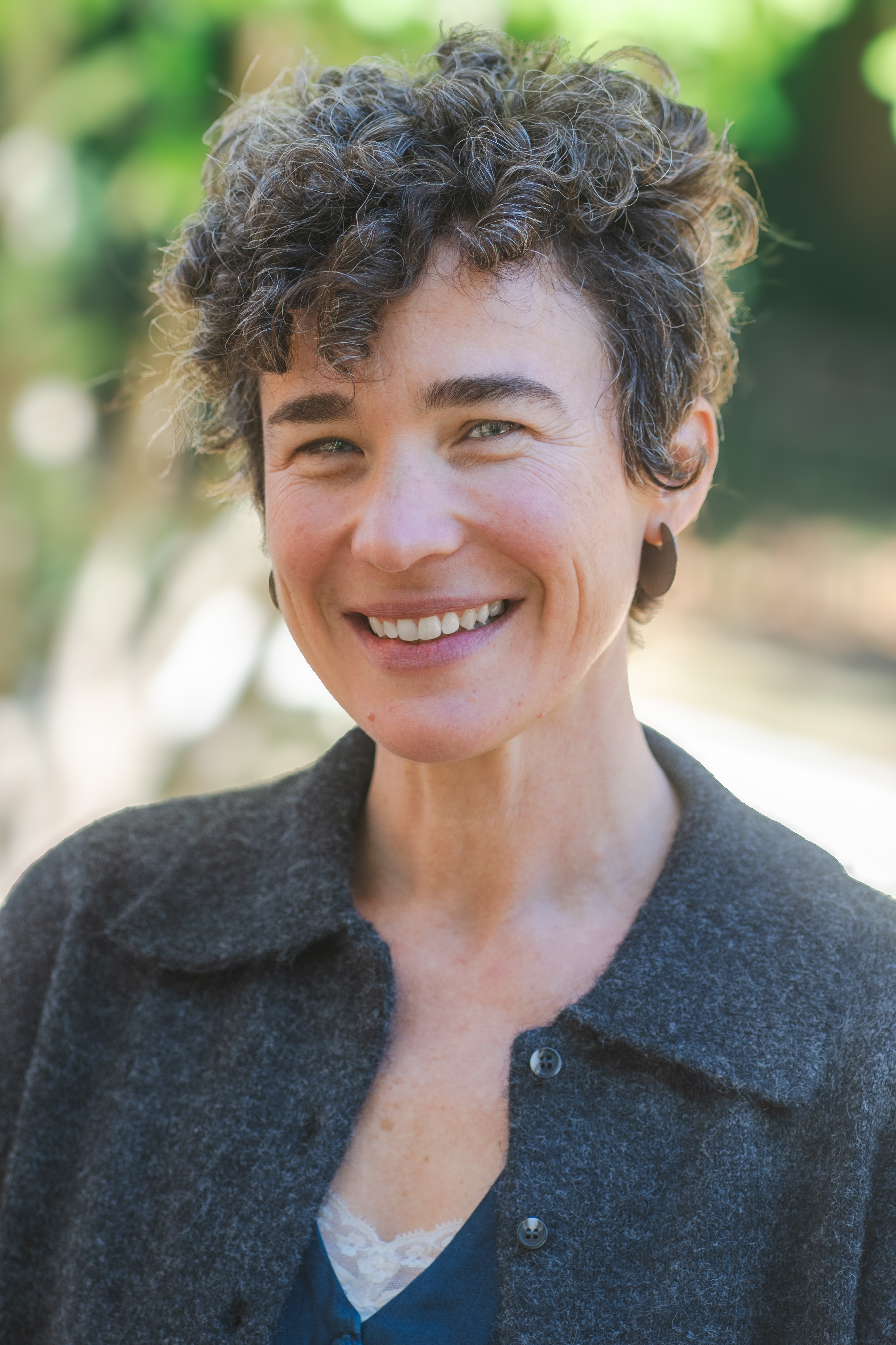 Portrait of Sarah Eichberger outdoors. Sarah is wearing a dark gray button-up jacket over a blue top. The background is softly blurred with green foliage and natural light.